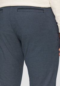 Dunkelblaue gemusterte Hose mit Karodesign, ausgestattet mit zwei Gesäßtaschen und dezenten Stickdetails. Glatte, leichte Stofftextur.