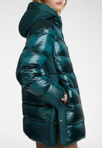 Turkooise donsjack met capuchon, voorzien van een glanzende, doorgestikte textuur en een knoopsluiting. Het ontwerp is boxy met een mid-lengte snit.