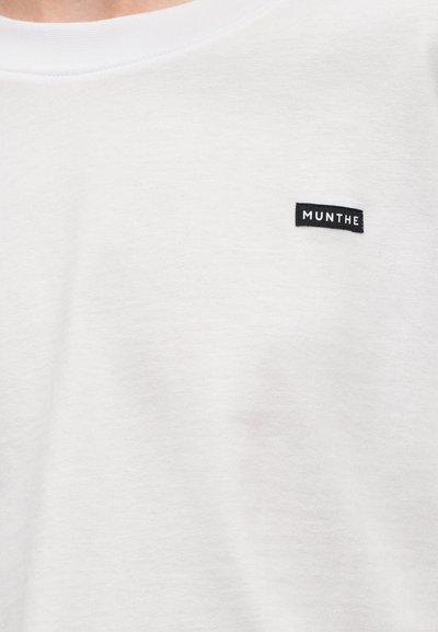 Λευκό βαμβακερό t-shirt με στρογγυλή λαιμόκοψη, λεία υφή και μια μικρή μαύρη ετικέτα με λογότυπο που αναγράφει "MUNTHE" στο μπροστινό μέρος.