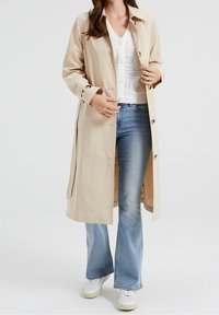 Beige lightweight trench coat, button-front design, long sleeves, side pockets; worn over white textured top and flared blue jeans.