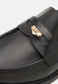 Black leather shoe with a smooth texture, featuring a decorative band and gold-tone hardware accent. Classic loafer design.