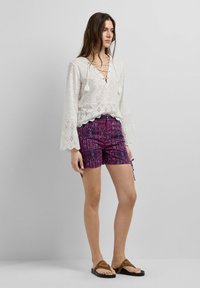 Žena stojí, má na sobě bílou halenku se šněrováním a dlouhými rukávy, fialové šortky s potiskem a hnědé sandály, na pozadí je jednolitá šedá zeď.