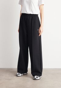 Pantaloni da jogging larghi neri con vita elastica, caratterizzati da un piccolo logo ricamato, abbinati a sneakers con motivo in bianco e nero.