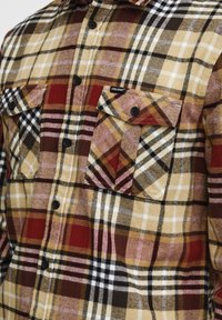 Camisa de franela con un diseño de cuadros en colores rojo, marrón, amarillo y blanco. Dos bolsillos en el pecho y botones negros. Textura suave.