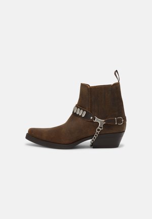 Bottine en daim marron avec bout pointu, talon bas empilé, chaîne argentée décorative et bride cloutée, avec languette à l'arrière.
