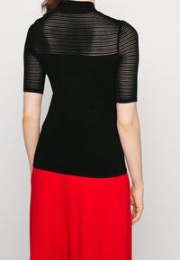 Černý pletený top s krátkými rukávy, který má průhledný pruhovaný design na horní části zad. Nosit s červenou sukní, která zvýrazňuje přiléhavý siluetu.