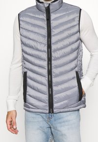 Grau gepolsterte Weste mit hohem Kragen, Reißverschluss, schwarzen Akzenten und Seitentaschen, ausgestattet mit einem strukturierten, gesteppten Design.