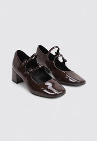 Par de zapatos Mary Jane de color marrón oscuro y brillante con bajos tacones en bloque y doble correa de hebilla sobre un fondo gris claro.
