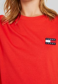 Rode katoenen T-shirt met een klein marineblauw en wit logo-patch met de tekst "TOMMY JEANS" op de linker borst. Korte mouwen.
