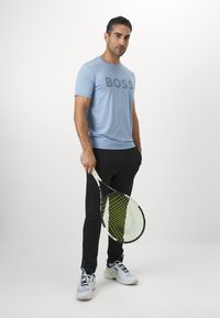 Camiseta de manga corta azul claro con el logotipo "BOSS", combinada con pantalones deportivos negros y zapatillas de tenis blancas, sosteniendo una raqueta de tenis.
