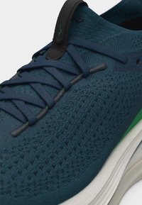 Κοντινό πλάνο μπλε πλεκτού αθλητικού παπουτσιού με σκούρα μπλε κορδόνια, μαύρο λεπτομέρεια στη γλώσσα και λευκή σόλα με πράσινες λεπτομέρειες.