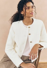 Giacca in corduroy bianco con scollo tondo e chiusure a pulsante dorato. Il modello indossa un top chiaro e pantaloni in tonalità neutra, seduto su una sedia di legno.