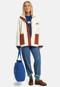 Crème fleece jack met bruine accenten, gedragen over een blauwe sweatshirt. Een blauwe tas, denim jeans, groene sokken en bruine schoenen maken de outfit compleet.