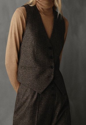 Person wearing a brown turtleneck sweater under a dark grey buttoned vest and matching pants, standing against a plain grey background.