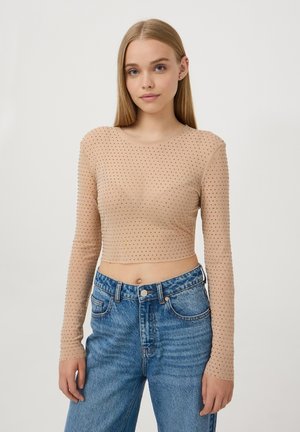 Camicia beige a maniche lunghe e taglio cropped con pois; materiale trasparente, scollatura rotonda, indossata con jeans in denim blu a vita alta.
