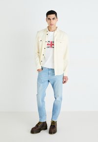 Camisa de botón color crema claro sobre una camiseta blanca con un gráfico de la bandera británica en rojo y azul, combinada con jeans azul claro y botas marrones.