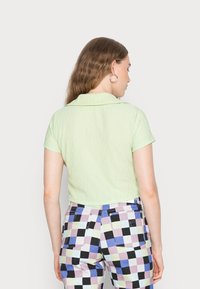 Camisa de manga curta verde-clara com colarinho, apresentando um tecido texturizado. Usada com calças xadrez multicoloridas em roxo, azul e preto.