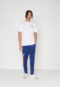 Jauns vīrietis ar pītu matu stilu stāv, valkājot baltu Adidas t-kreklu, zilas sporta bikses ar sarkaniem un gaiši zilajiem rakstiem, kā arī baltas sporta kurpes.