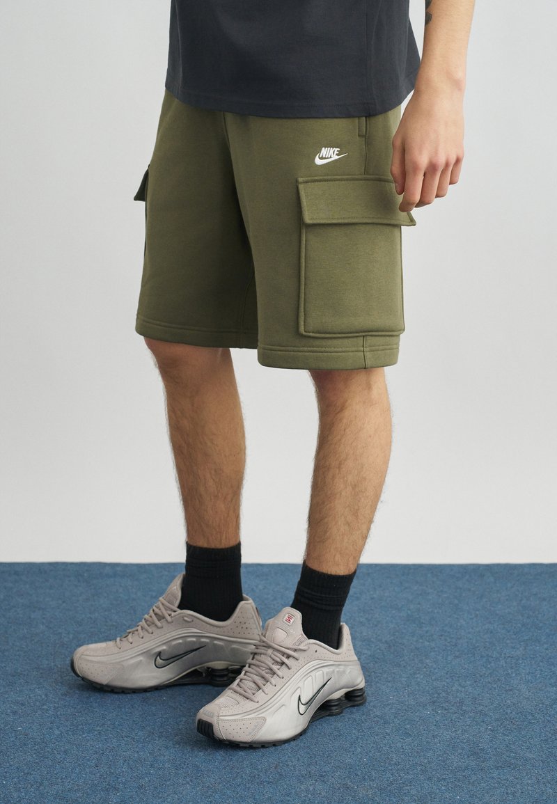 Άτομο που φοράει λαδί cargo σορτς της Nike, μαύρες κάλτσες και ανοιχτό γκρι αθλητικά παπούτσια Nike, στέκεται σε μπλε χαλί.