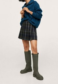 Groene rubberen laarzen tot de knie, met ribbelzool, gecombineerd met een blauwe gebreide trui en een geruite geplisseerde rok in verschillende tinten marineblauw en groen.