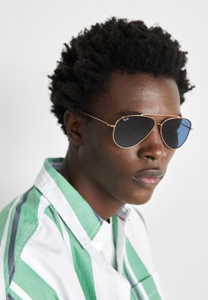 Pilotensonnenbrille mit dunklen Gläsern, goldenen Metallrahmen und dünnen Bügeln. Die Person trägt ein grün-weiß gestreiftes Hemd.