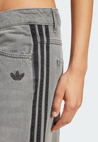 Pantalones de mezclilla gris con tres franjas laterales negras, un pequeño logo negro en el bolsillo y un acabado texturizado. Se pueden llevar de forma casual.