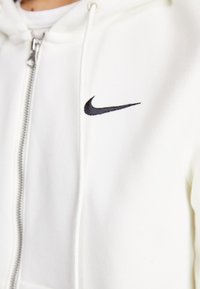 Sweatshirt branco com fecho éclair, feito de tecido macio, apresentando um logo da Nike bordado a preto no lado esquerdo do peito e capuz com cordão.