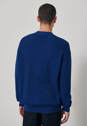 Hombre con cabello corto y oscuro que lleva un suéter de punto acanalado azul y pantalones negros, de espaldas sobre un fondo claro y liso.