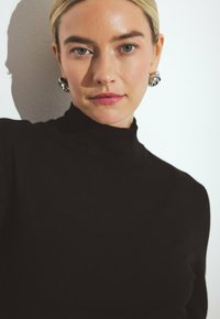 Jersey de cuello alto negro con un diseño ajustado. La modelo lleva pendientes de plata, mostrando una textura suave y una silueta minimalista.