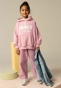 Różowa bluza z kapturem z nadrukiem "DANCE Studio", prążkowane różowe spodnie oraz dżinsowa kurtka trzymana w ręku. Dodatki obejmują białe trampki. Gładkie tekstury.