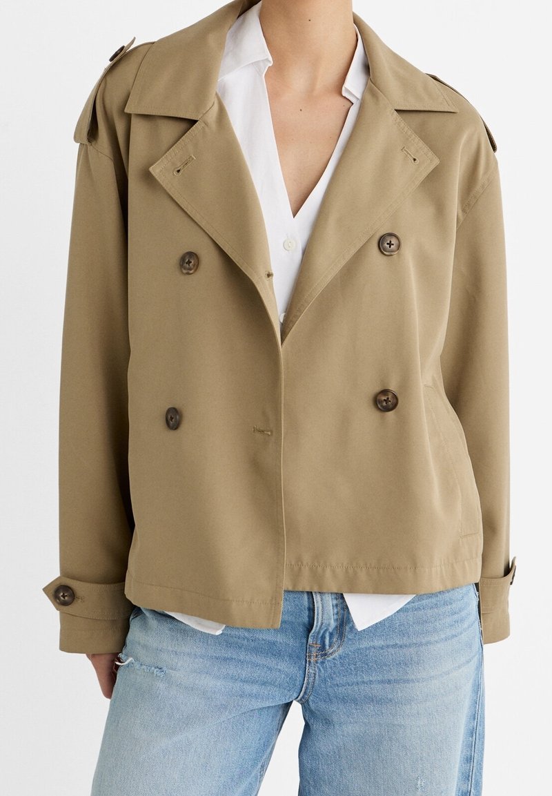 Beige double-breasted jacket with large brown buttons, wide lapels, and shoulder epaulets, layered over a white shirt. Cotton-blend fabric.