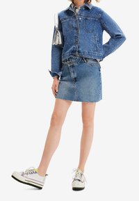 Kvinna som bär en blå jeansjacka med små färgglada prickar, en jeanskjol med fransar, vita plattformssneakers och bär en silverhandväska.