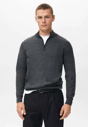 Sudadera gris oscuro con cremallera hecha de una tela suave, que presenta un cuello alto y mangas largas, llevada sobre una camisa blanca y combinada con pantalones negros.