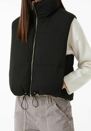 Femme portant un gilet matelassé sans manches noir avec fermeture éclair et cordons, un haut blanc à manches longues et un pantalon rayé, les mains dans les poches.