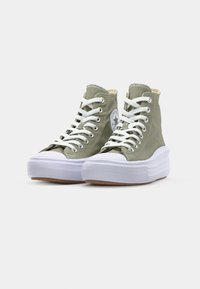 Sneaker alte in tela verde oliva con lacci bianchi, suola in gomma bianca spessa e una patch circolare del logo sul lato.