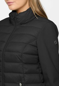 Woman wearing black quilted puffer jacket with zip-up front and smooth fabric sleeves, visible brand logo on upper left sleeve.