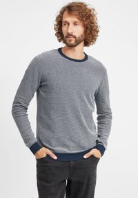 Maglione grigio a maglia texturizzata con scollatura rotonda e polsini e orlo a costine blu navy. Indossato sopra jeans scuri, presenta un motivo sottile.