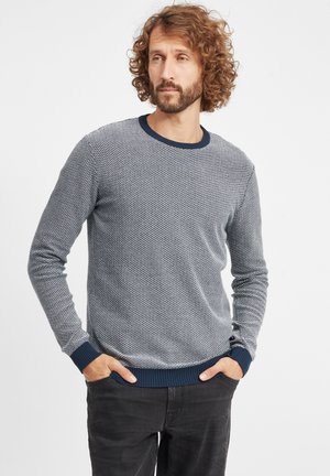 Gray, textured knit sweater with a crew neckline and navy ribbed cuffs and hem. Worn over dark jeans, features a subtle pattern.
