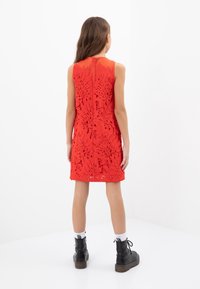 El vestido rojo sin mangas presenta intrincados patrones de flores cortados; cierre de cremallera en la parte posterior, combinado con botas negras hasta el tobillo y calcetas blancas.