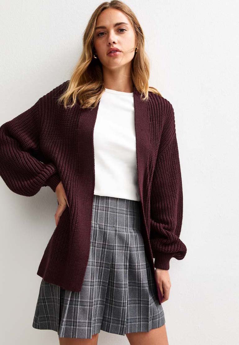 Cardigan in magenta lavorato a maglia con vestibilità ampia, maniche larghe e texture a coste, indossato sopra una maglietta bianca e una gonna plissettata a quadri grigi.