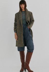 Brun hørslagsfrakke over en teal-top og marineblå bukser, parret med brune højhælede støvler; bemærkelsesværdige teksturer og afslappet pasform.