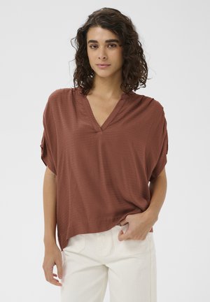 Femme aux cheveux bouclés portant un haut ample couleur rouille à col en V et un pantalon blanc, debout avec une main dans la poche contre un fond blanc.