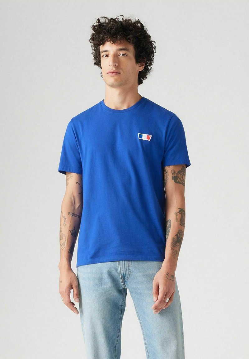 Hombre con cabello rizado que lleva una camiseta azul con un pequeño parche rojo, blanco y azul, combinado con unos jeans de mezquita claros frente a un fondo liso.