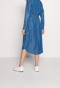 Abito camicia in denim con vita cintata, maniche lunghe e orlo curvo, in una tonalità di blu medio; abbinato a sneaker bianche.