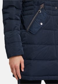 Marineblauwe pufferjas met gequilt textuur, voorzien van een ritssluiting op de borst en twee knopen; gladde stof met zachte isolatie.
