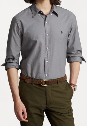 Hombre con camisa gris abotonada con mangas remangadas, cinturón marrón, pantalones verde oliva y un reloj de pulsera en la muñeca izquierda.
