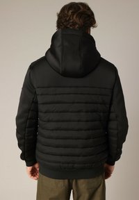 Homme portant une veste à capuche noire rembourrée et un pantalon vert olive, se tenant de dos à la caméra sur un fond uni.