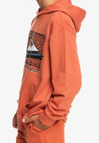 Sudadera naranja hecha de tejido suave, con un estampado gráfico en la parte frontal que presenta imágenes de montañas y un logotipo, y con un bolsillo tipo canguro.