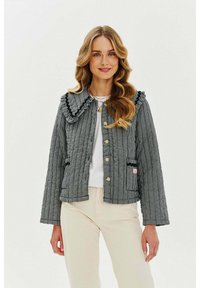 Prošívaná černobílá kostkovaná bunda s velkým řaseným límcem, zlatými knoflíky a předními kapsami; nošena přes bílý top a béžové kalhoty.