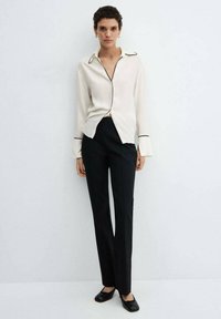 Cremfarbene Bluse mit schwarzer Paspelierung, langen Ärmeln und einem lässigen Schnitt, kombiniert mit eng anliegenden schwarzen Hosen. Das Model trägt schwarze Ballerinas.